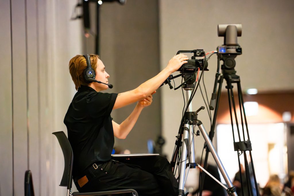 An Audio Visual Technician wearing a headset adjusts a video camera on a tripod in an indoor setting, possibly at an event or conference.