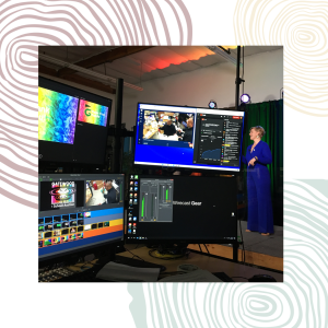 In the background, a woman stands on a stage. In the foreground, large screens and monitors display a workstation for audio visual technicians.