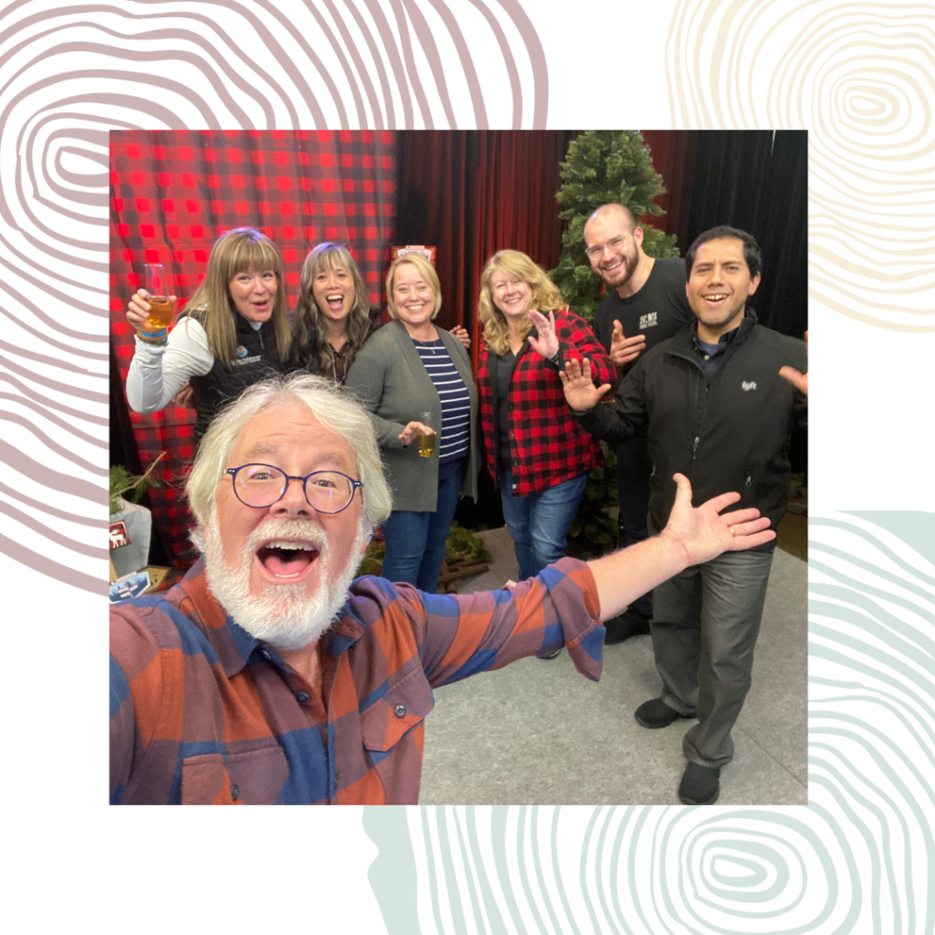 A man with white hair and beard stands with his arm outstretched to a group of six people behind him.