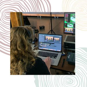 A woman with her back turned towards us wears headphones and works at a laptop