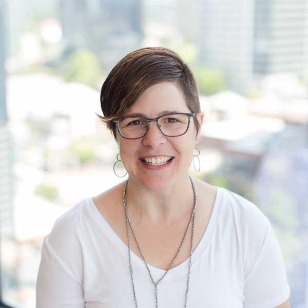 A person with short brown hair, glasses, and a white shirt smiles at the camera with a blurred cityscape in the background, embodying the professional spirit of Seattle AV Services for Hotels and Venues.