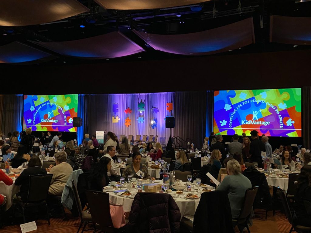 A large banquet hall with round tables full of people dining; two screens display the KidVantage logo and colorful puzzle piece graphics on stage.