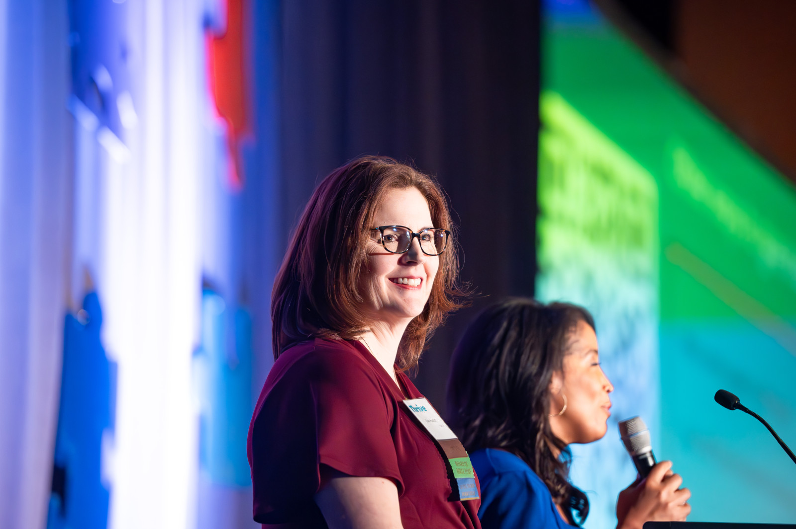 LB5_0090-Enhanced-NR Two women stand on stage at a conference—one smiles at the audience while the other speaks into a microphone, supported by Seattle audio visual services for auctions and fundraisers.