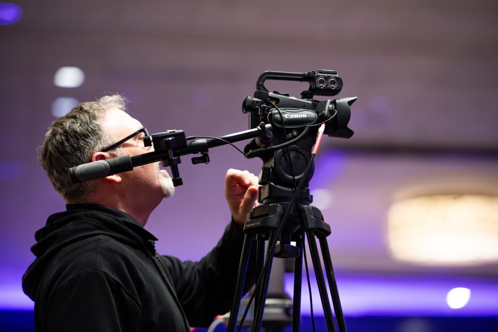 A man in glasses operates a Canon video camera mounted on a tripod indoors, adjusting the camera settings.