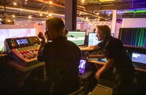 Two AV technicians sit in a sound booth using a digital sound mixer.