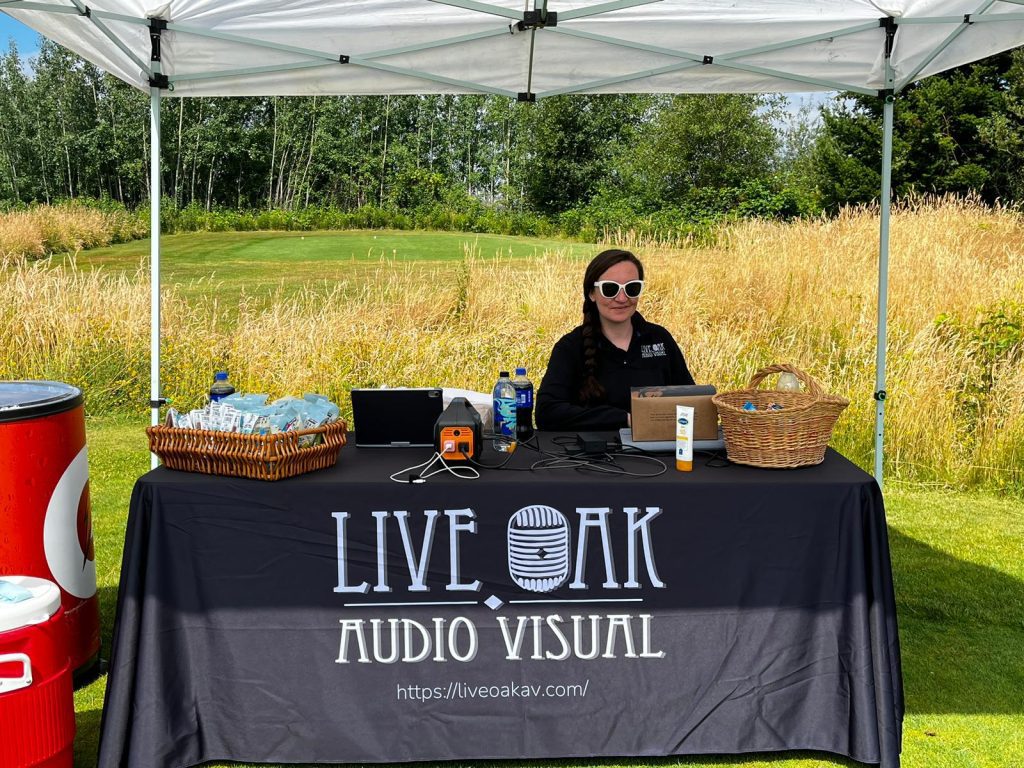 A woman sits behind a table covered with a "Live Oak Audio Visual" cloth under a canopy on a grassy field, with electronic equipment, snacks, and sunscreen on the table. Contact Live Oak AV for your event needs.