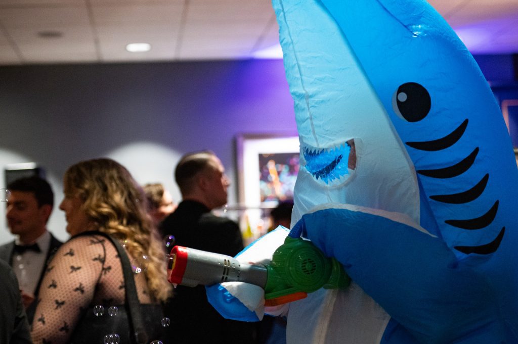 Person in a blue inflatable shark costume holding a toy water gun at an indoor event, with guests in formal attire and Seattle audio visual services for auctions and fundraisers enhancing the atmosphere in the background.