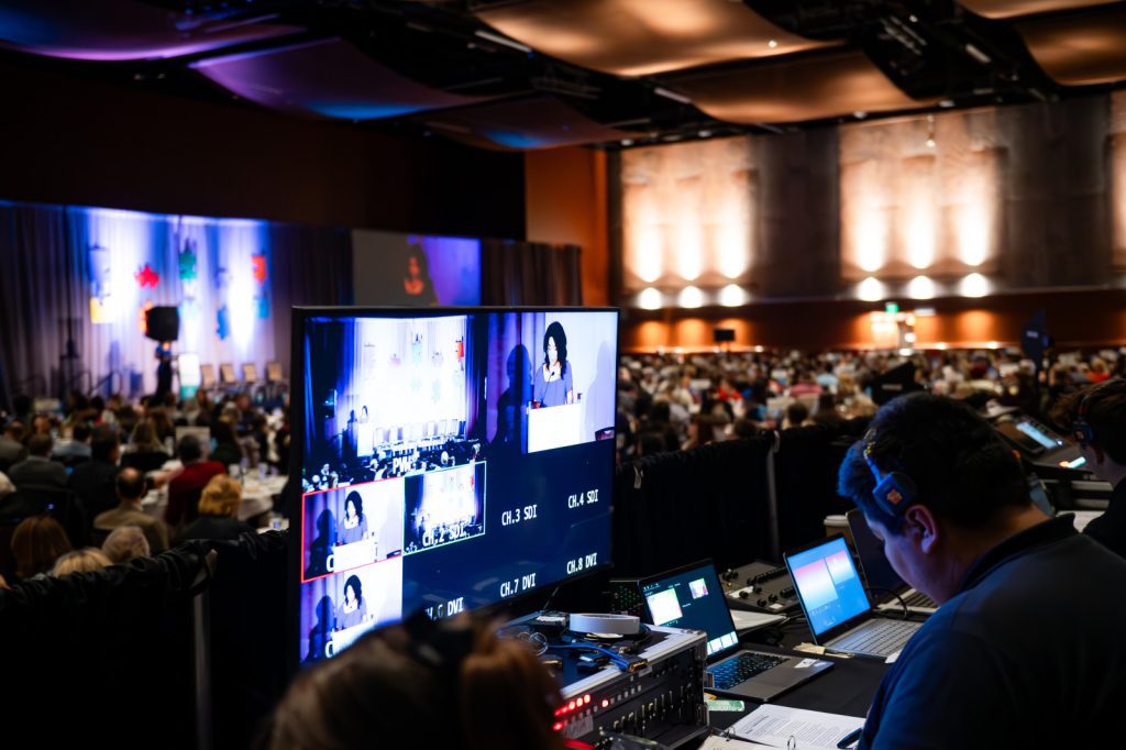 A technician with Production Partners operates audio-visual equipment at the back of a large conference room, with multiple screens showing a speaker addressing an audience.