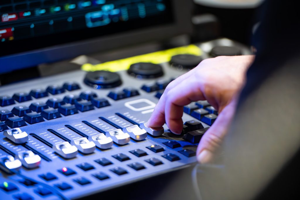 A hand adjusts sliders on a professional audio mixing console, with a computer monitor and additional controls visible in the background.