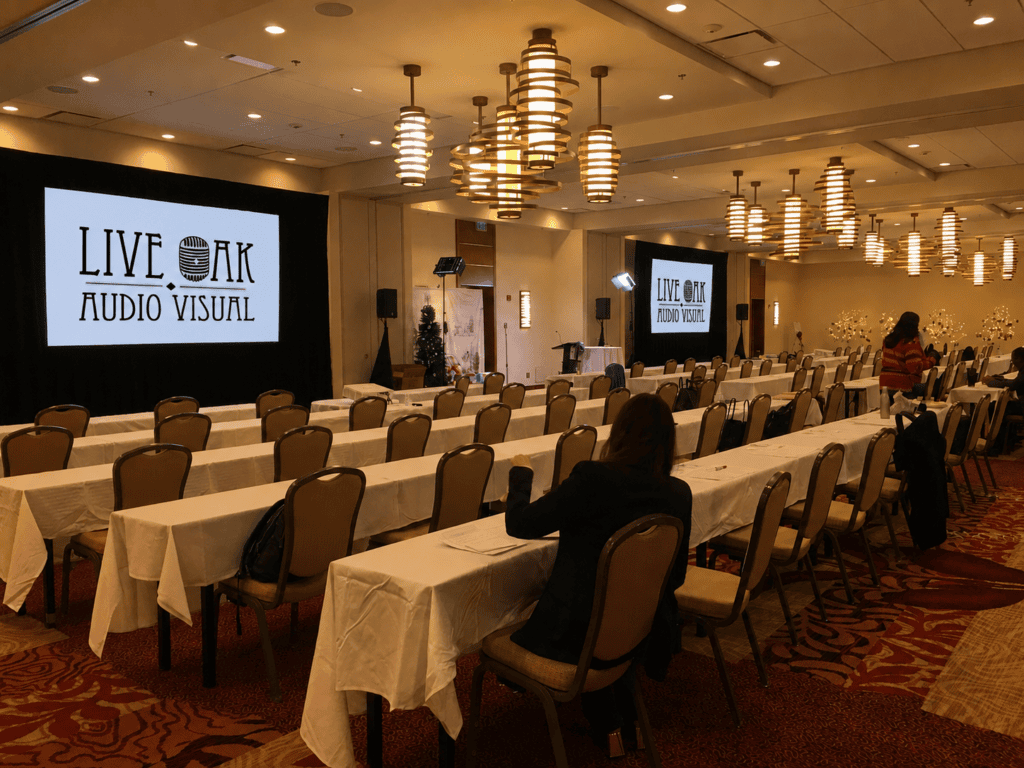Image of a conference room set up for a speaking event, with screens featuring the Live Oak AV logo.