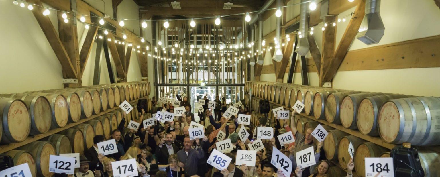 A crowd inside a barrel-lined room holds up numbered signs, as if participating in an auction or bidding event under string lights—perfect inspiration for your next auction planning or virtual auction experience.