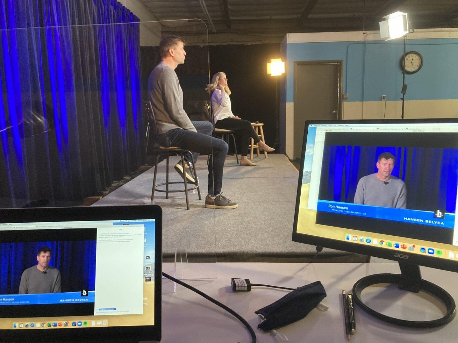 A man and woman sit on a stage with blue curtains, preparing for an event as part of conference planning, while a live video feed is displayed on two monitors in the foreground. Studio lights and a clock are visible.