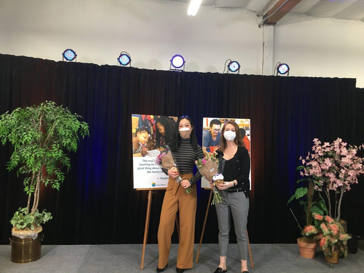 Two people wearing masks stand on a stage holding bouquets, in front of a poster with photos and text. The stage, decorated with plants and spotlights overhead, celebrates lessons learned from virtual auctions.