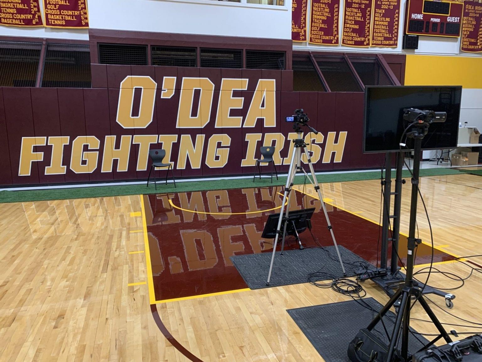 A gymnasium with "O'DEA FIGHTING IRISH" on the wall, two empty chairs, cameras, lights, and a screen set up on the polished wooden floor—perfect for virtual graduation ceremonies or innovative graduation ideas.