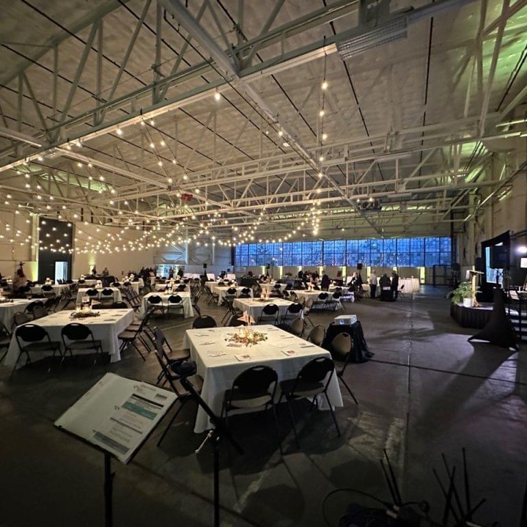 Large indoor event space with string lights, round and square tables set for guests, and a few people scattered throughout the room—ideal for auctions or fundraisers with professional Seattle audio visual services.