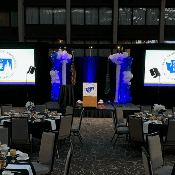 A banquet hall is set up for corporate events with round tables, a podium, and two large screens displaying the Pemiscot County School Board Association logo.