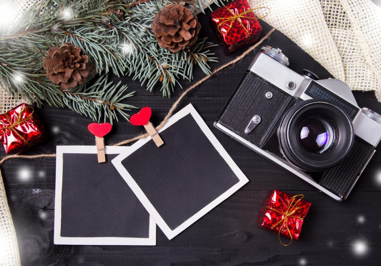 A vintage camera, blank photo frames, pine branches with cones, and small wrapped gifts are arranged on a dark wooden surface.