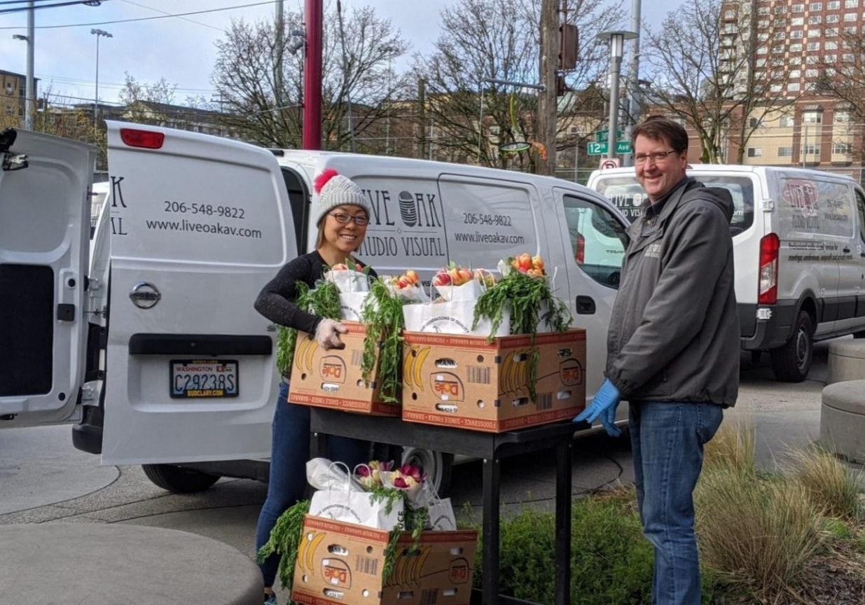 Two people stand by flower-filled boxes on a cart outdoors, next to white delivery vans with "Live Oak AV" branding.