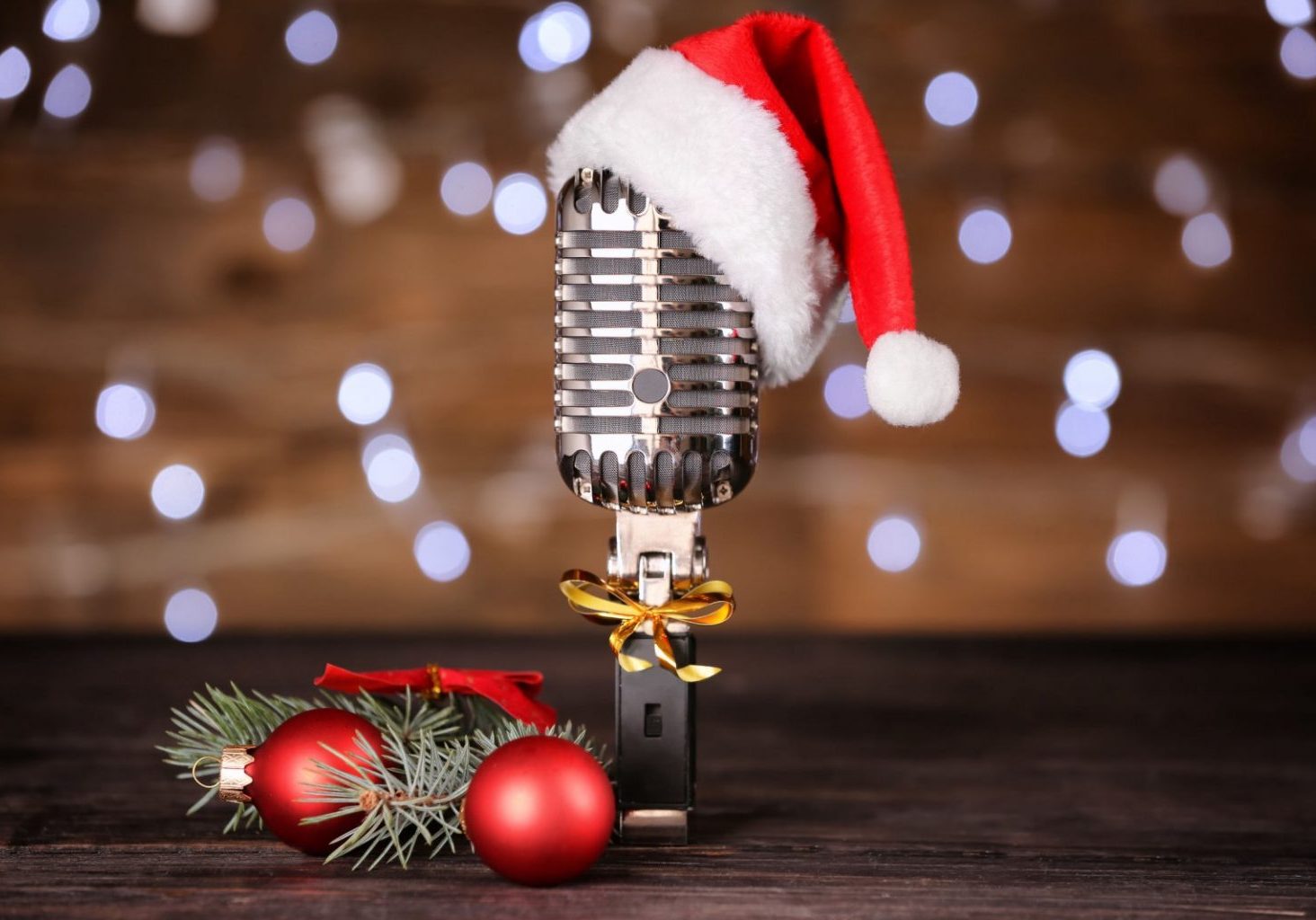 A vintage microphone with a Santa hat, gold ribbon, and red Christmas ornaments with pine branches, set against a blurred background of white lights—perfect for your Tech Gift Guide this holiday season.