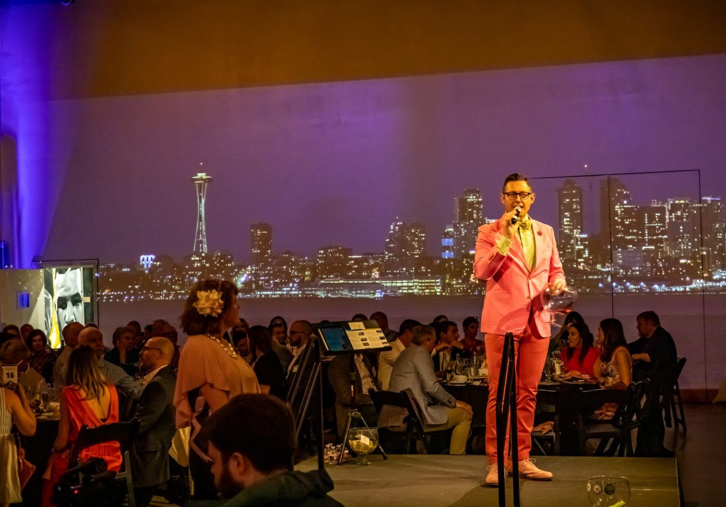 Blog main image A man in a pink suit speaks into a microphone on stage at an indoor event, with a projected city skyline in the background and seated guests watching.