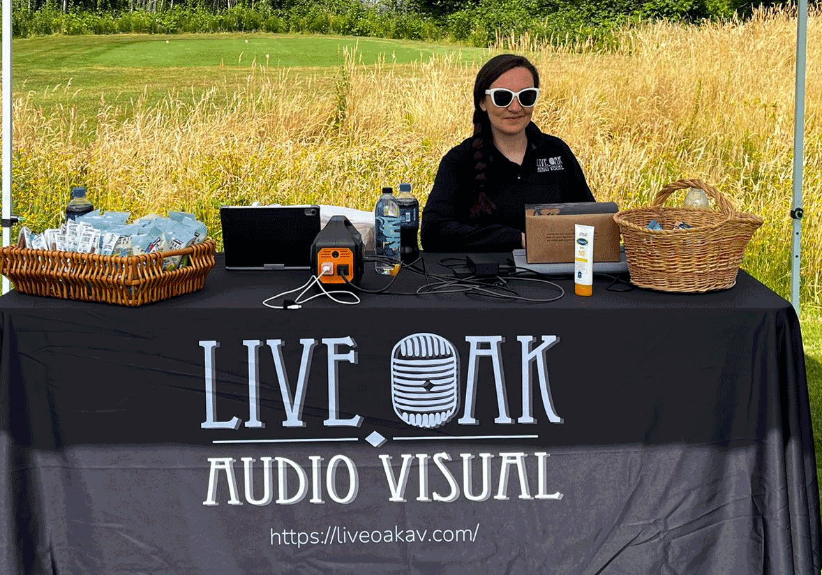 A woman wearing sunglasses sits behind a table with audio visual equipment, snacks, and baskets under a tent with a "Live Oak Audio Visual" tablecloth. Contact Live Oak AV for your event’s audio visual needs.