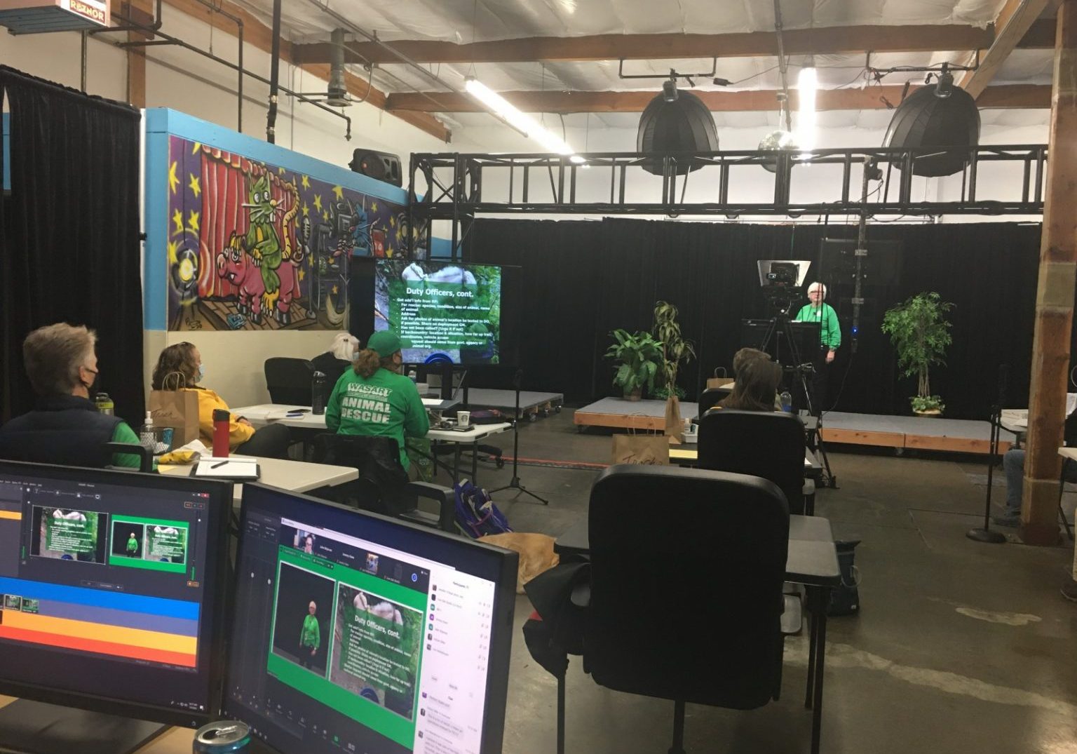 People sit in a studio with computers and monitors as a presenter speaks at a podium. Screens display environmental topics, supporting immersive training in a space featuring plants and a colorful mural.