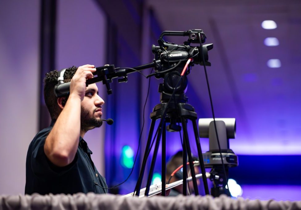 A man wearing a headset operates a professional video camera on a tripod in a conference room with blue and purple lighting, showcasing uncomplicated AV services for Seattle events.