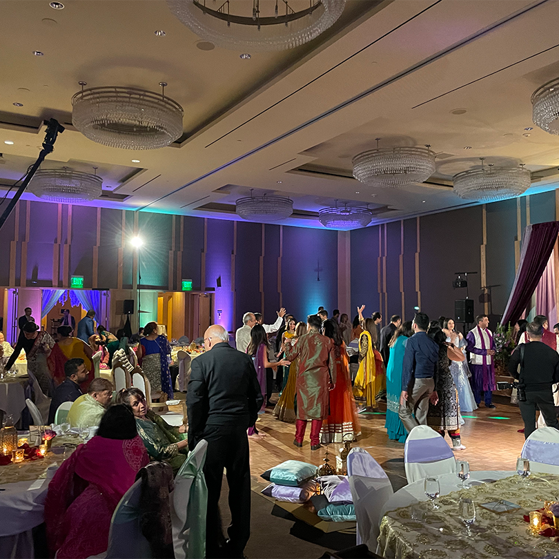 Copy-of-Nadim-Laiwala-Wedding-11.19.21-PC-Ryan-Keyes_Dancing-3 People dressed in formal and traditional attire dance and socialize in a decorated banquet hall, enhanced by AV services for Seattle weddings and private events, with tables, draped chairs, and sparkling chandeliers.