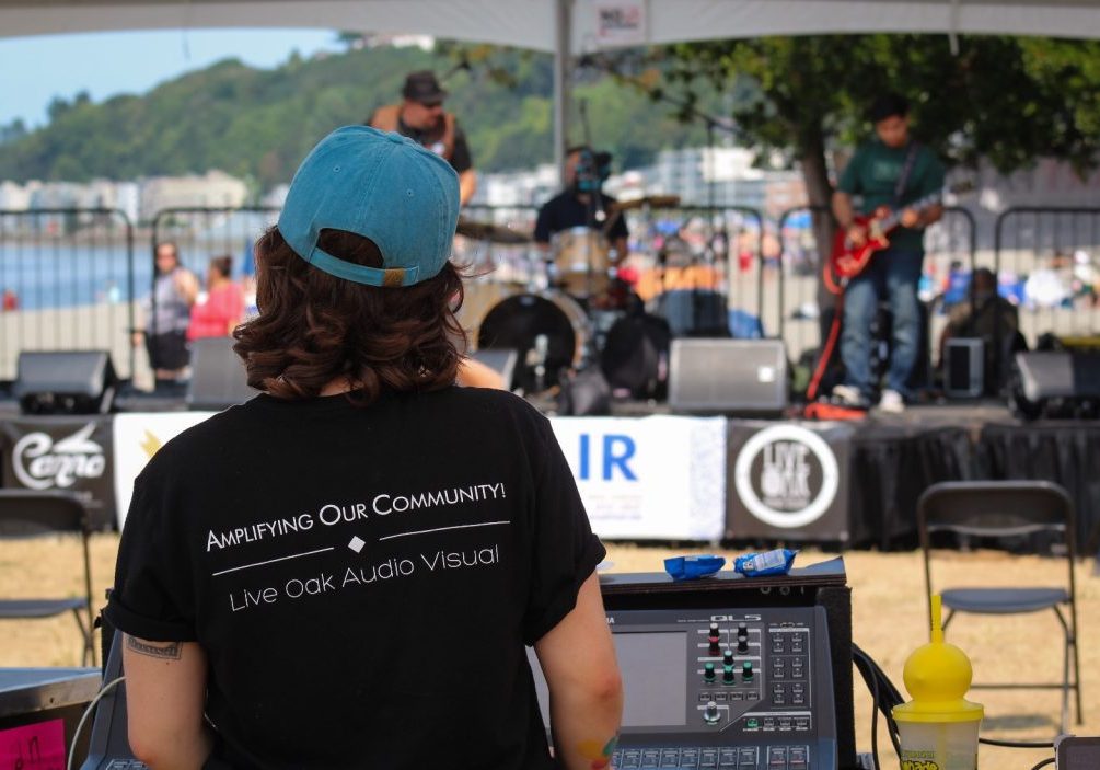 Copy of WhatsApp Image 2024-07-24 at 10.01.27 A person operates sound equipment at an outdoor concert, facing a stage with a band performing; people and a body of water are visible in the background, highlighting uncomplicated AV services for Seattle events.