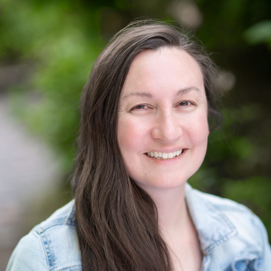 Crystal Cliff HS 2023 A woman with long brown hair and a light denim jacket, part of the Above-and-Beyond Client Experience Team, smiles outdoors against a blurred green background.