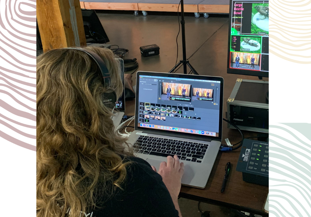 A woman with her back turned towards us wears headphones and works at a laptop