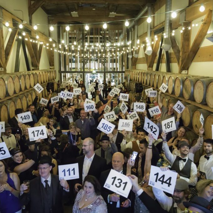 Fundraisers second photo middle A large group of people at an indoor auction hold up numbered paddles, surrounded by wine barrels and string lights overhead, with Seattle audio visual services for auctions and fundraisers enhancing the vibrant atmosphere.