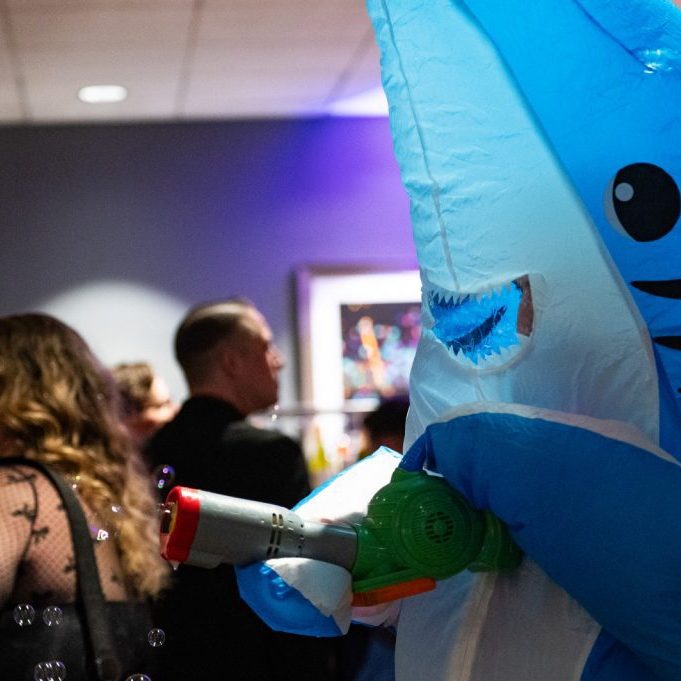 Fundraisers third image right Person in a blue inflatable shark costume holding a toy water gun at an indoor event, with guests in formal attire and Seattle audio visual services for auctions and fundraisers enhancing the atmosphere in the background.