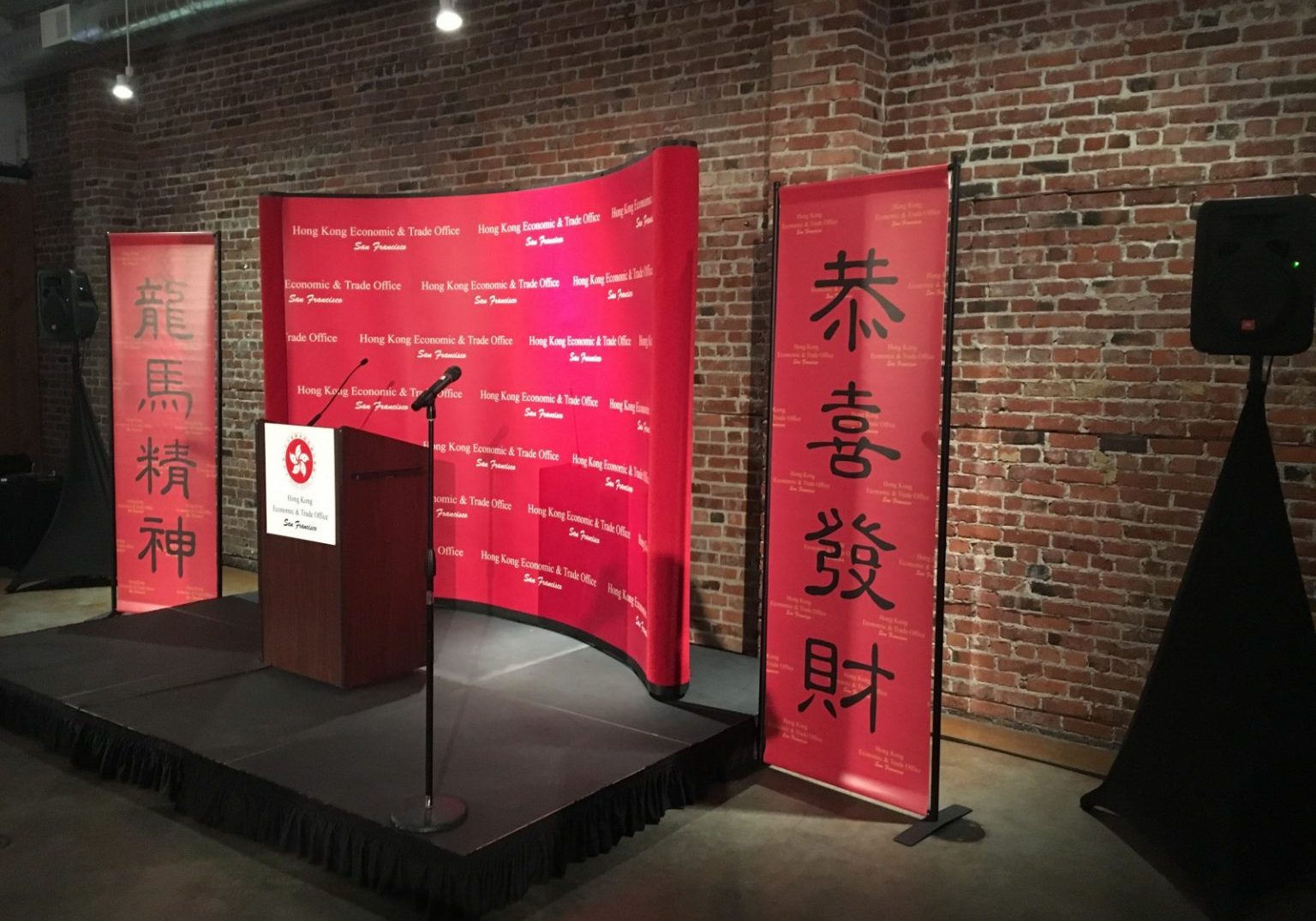 A podium with microphones stands on a small stage, flanked by red banners with Chinese characters, set against a brick wall background—creating an inclusive space ideal for a hybrid event that fosters equality and connection.