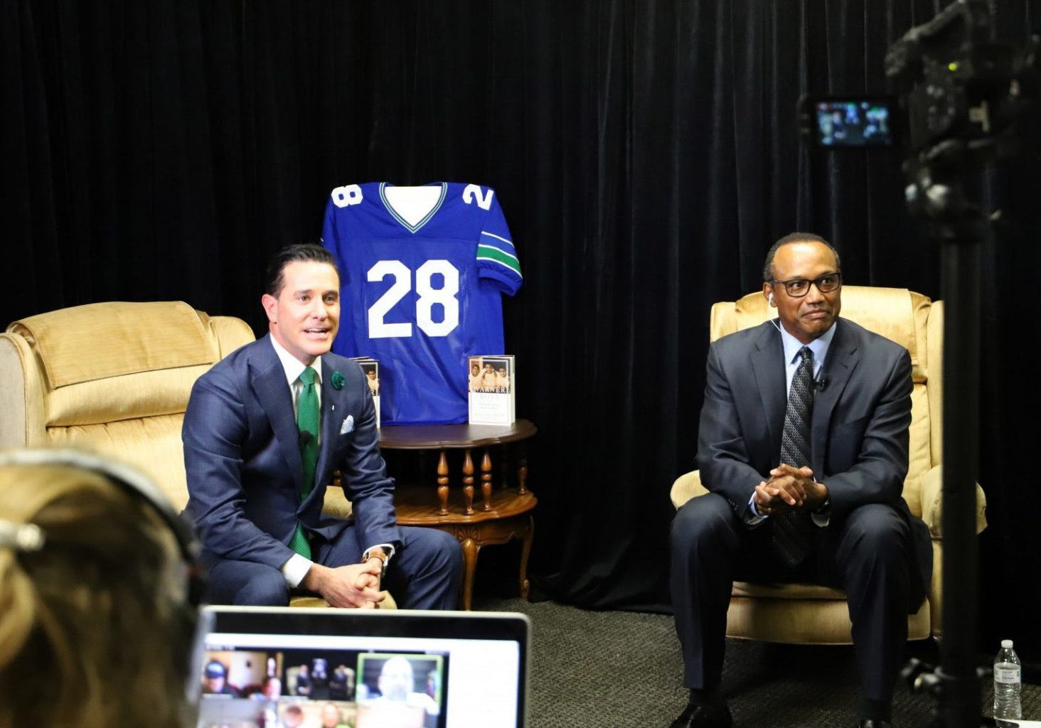 Two men in suits sit on armchairs in a studio with a football jersey displayed behind them, discussing creative holiday party ideas. A camera and laptop are visible in the foreground.