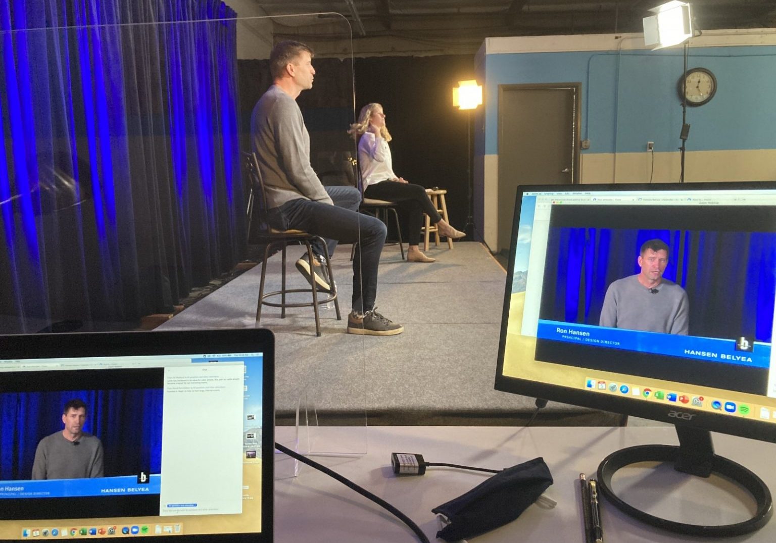A man and woman sit on a stage with blue curtains, preparing for an event as part of conference planning, while a live video feed is displayed on two monitors in the foreground. Studio lights and a clock are visible.