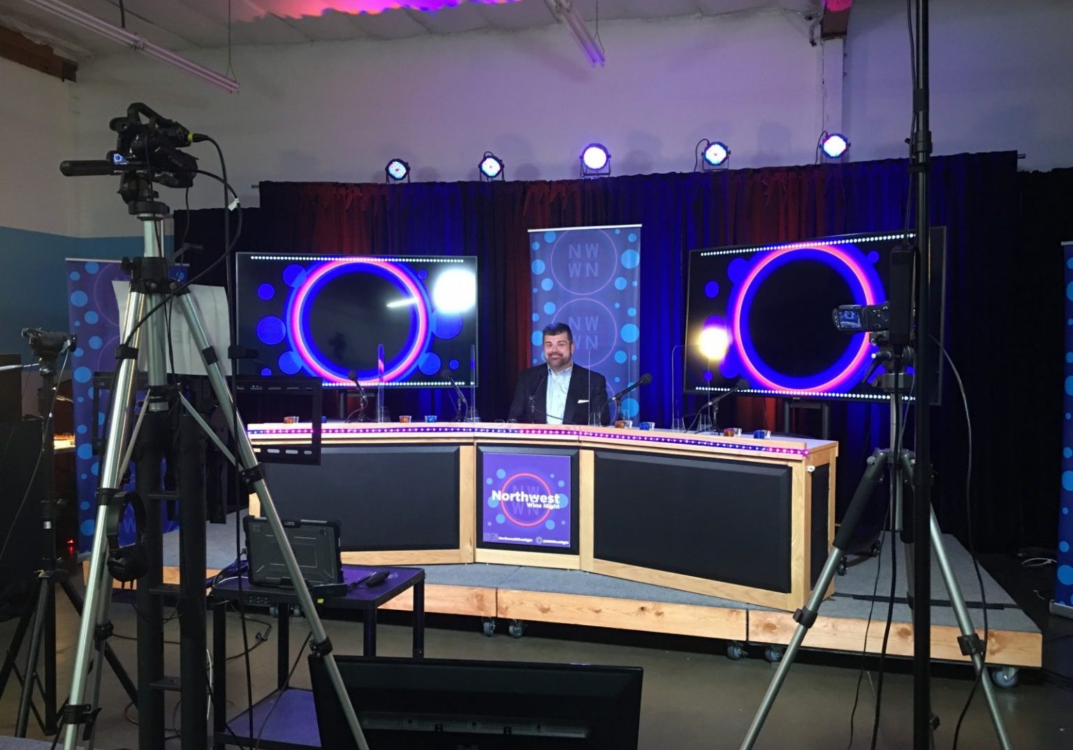 A man sits at a news desk in a studio with cameras, lights, and monitors displaying circular graphics behind him, ready to discuss the latest on Northwest Wine Night and the wine industry’s COVID pivot.