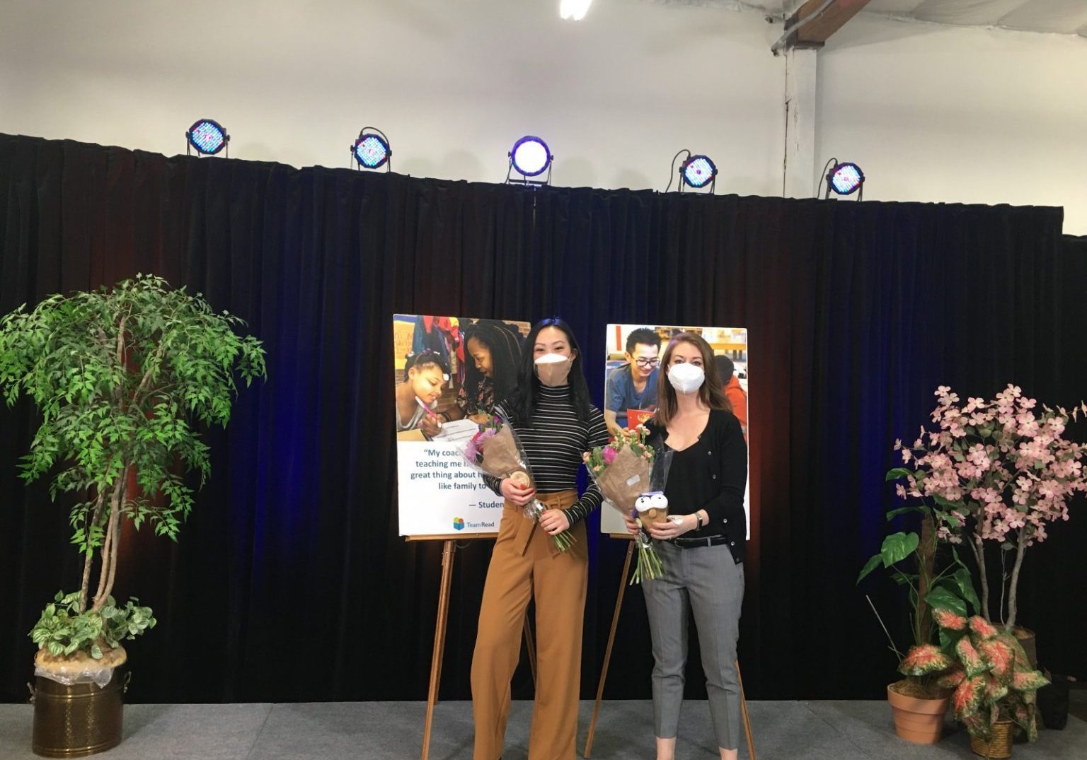 Two people wearing masks stand on a stage holding bouquets, in front of a poster with photos and text. The stage, decorated with plants and spotlights overhead, celebrates lessons learned from virtual auctions.
