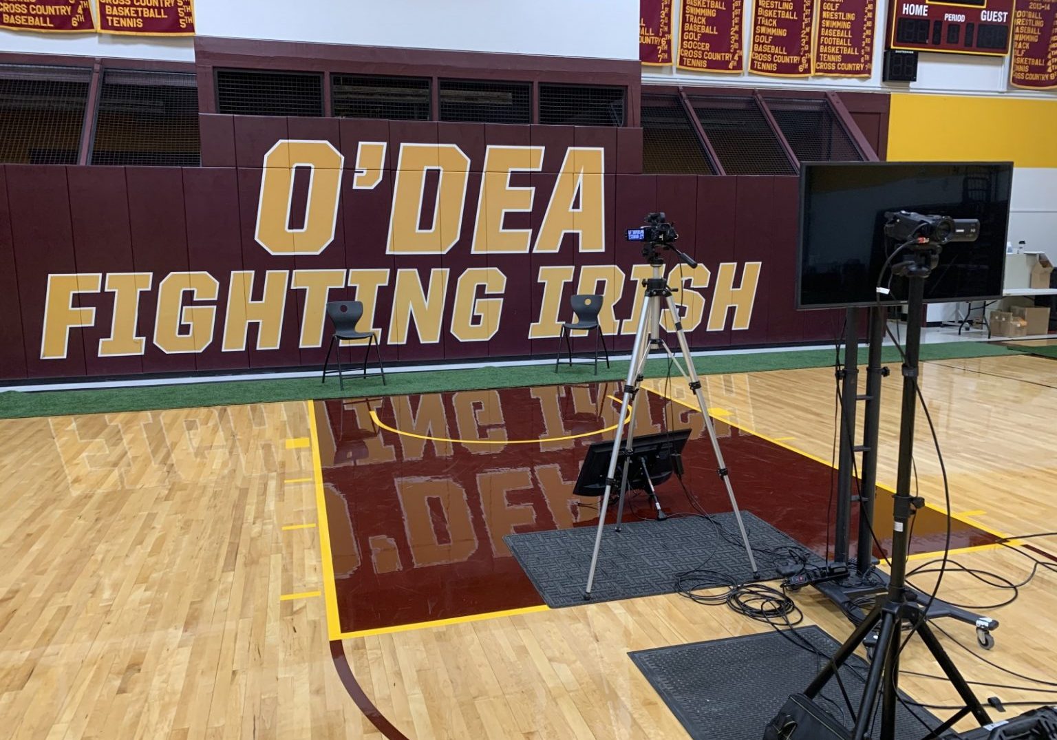 A gymnasium with "O'DEA FIGHTING IRISH" on the wall, two empty chairs, cameras, lights, and a screen set up on the polished wooden floor—perfect for virtual graduation ceremonies or innovative graduation ideas.