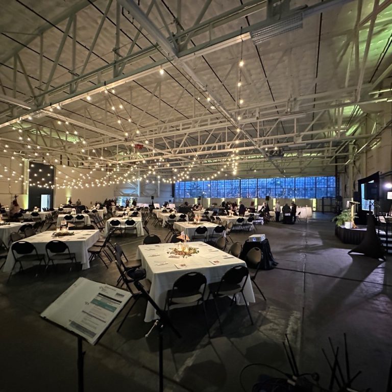 Large indoor event space with string lights, round and square tables set for guests, and a few people scattered throughout the room—ideal for auctions or fundraisers with professional Seattle audio visual services.
