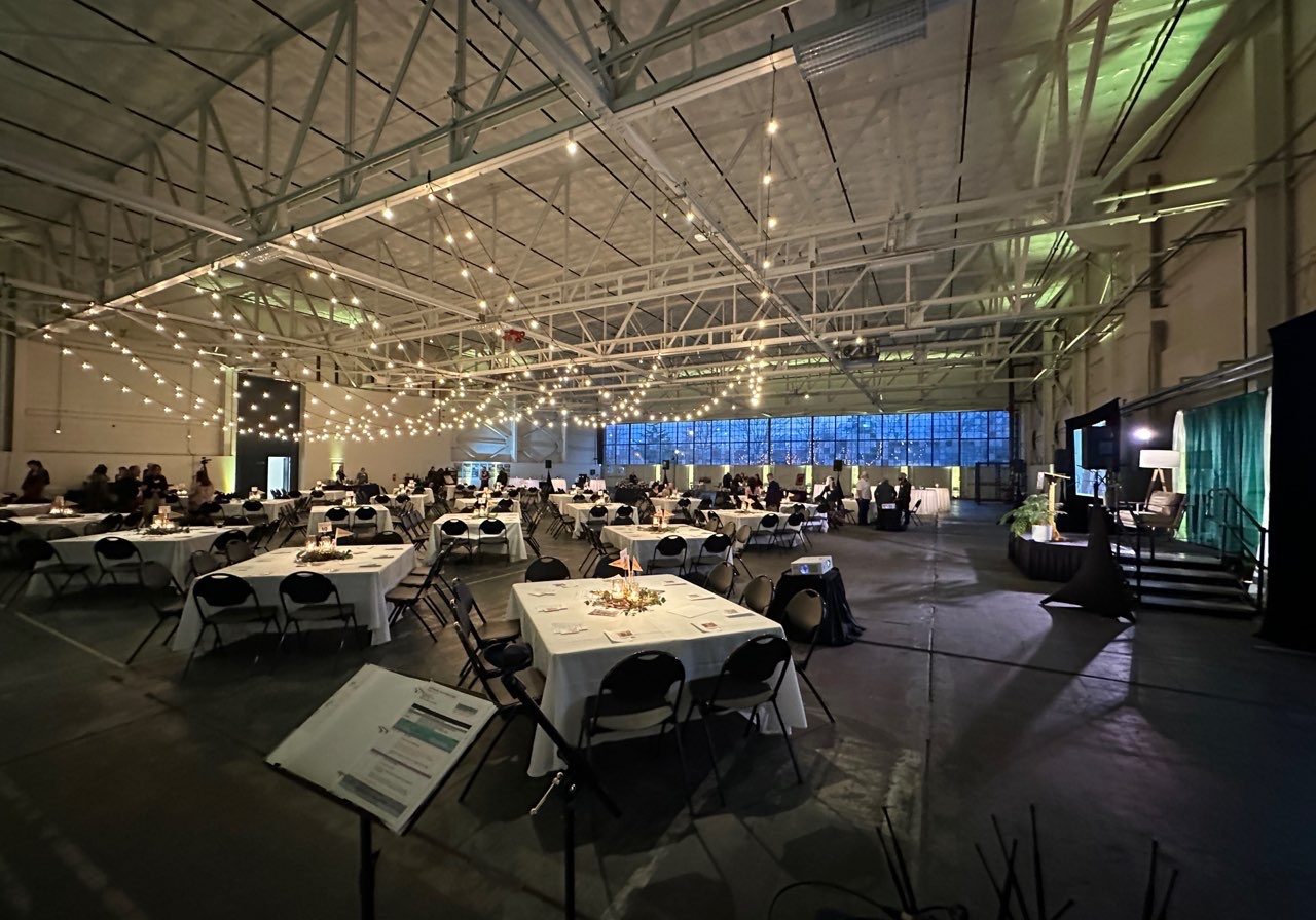 Large indoor event space with string lights, round and square tables set for guests, and a few people scattered throughout the room—ideal for auctions or fundraisers with professional Seattle audio visual services.