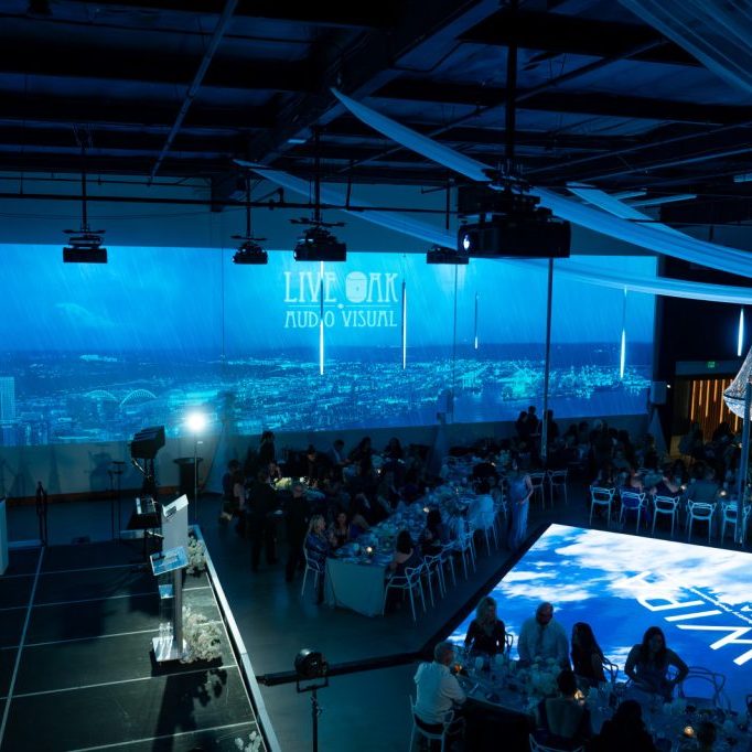A large banquet hall with blue lighting, projection screens displaying "LIVE ONLY AUDIO VISUAL," guests seated at tables, and a stage area with musical instruments.