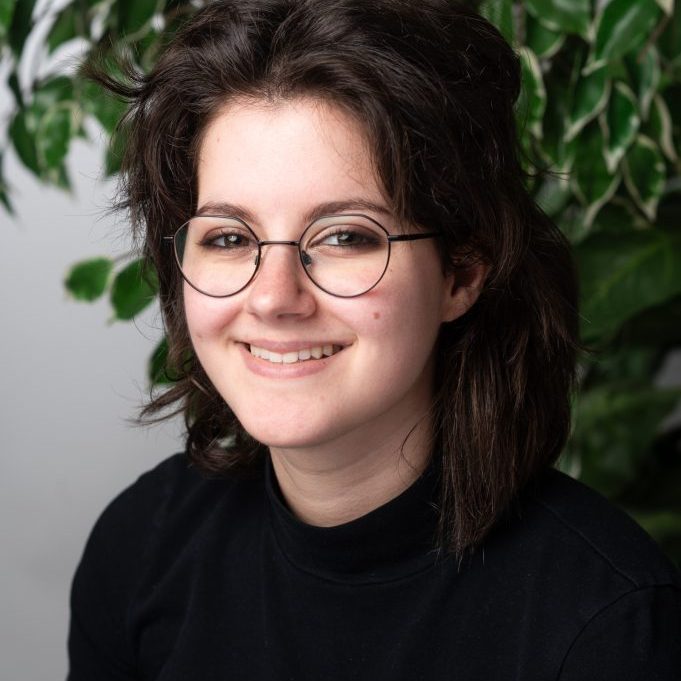 Jess Makler HS 2024 A person with dark, wavy hair and glasses, wearing a black turtleneck, smiles in front of a leafy green plant—embodying the friendly spirit of the Above-and-Beyond Client Experience Team.