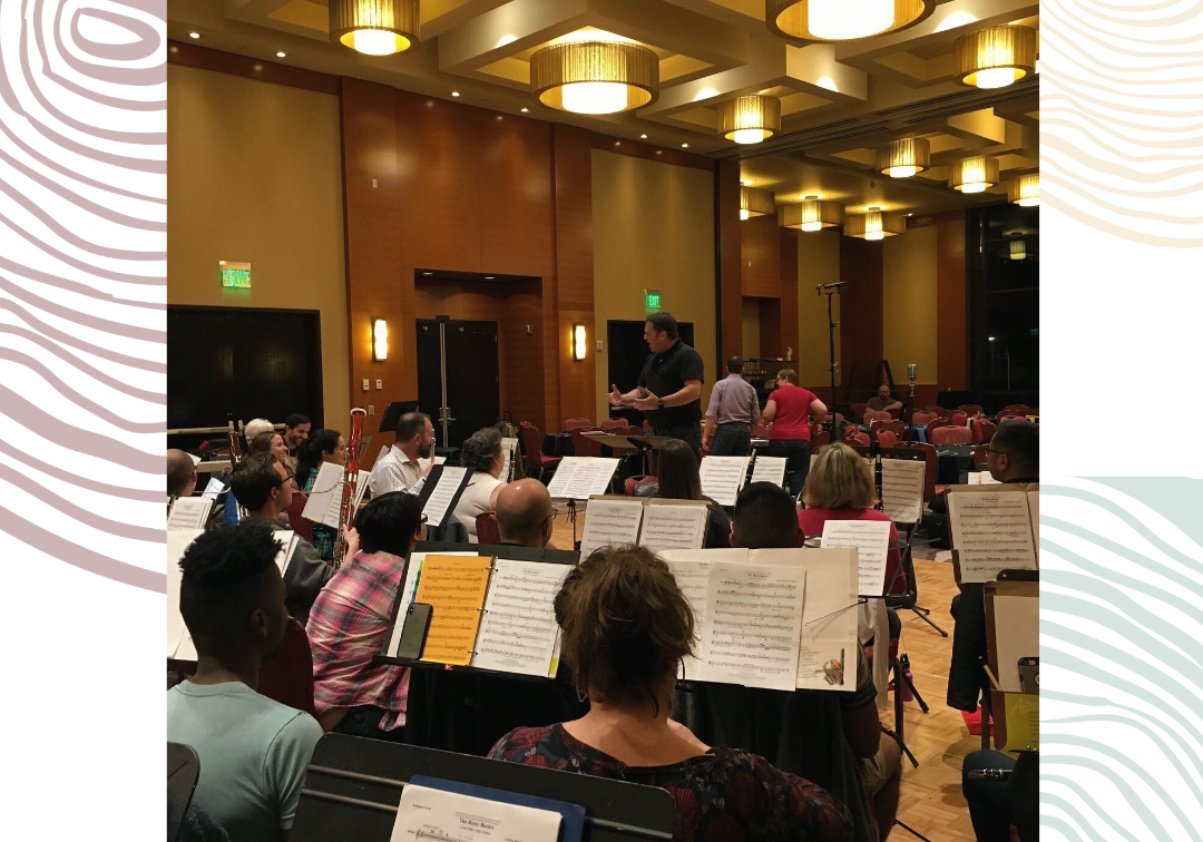 A conductor stands before a seated orchestra in a well-lit rehearsal room in Rainbow City, with musicians holding instruments and sheet music on stands, creating a safe harbor for the LGBTQIA+ community.