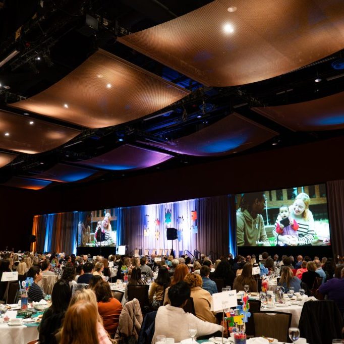 Large conference room filled with people seated at round tables, facing a stage equipped with large Audio Visual screens displaying a video of two people talking.