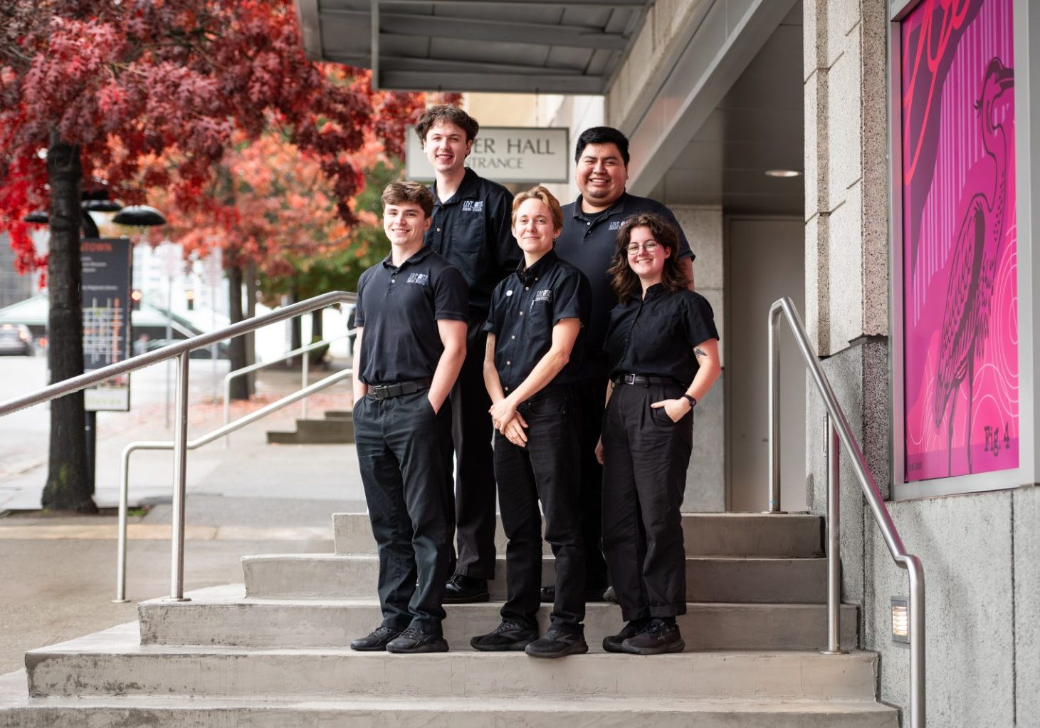 Five members of the Above-and-Beyond Client Experience Team, dressed in black uniforms, stand in a line on outdoor steps beside a building, with autumn trees and a sign reading "Fisher Hall Entrance" visible in the background.