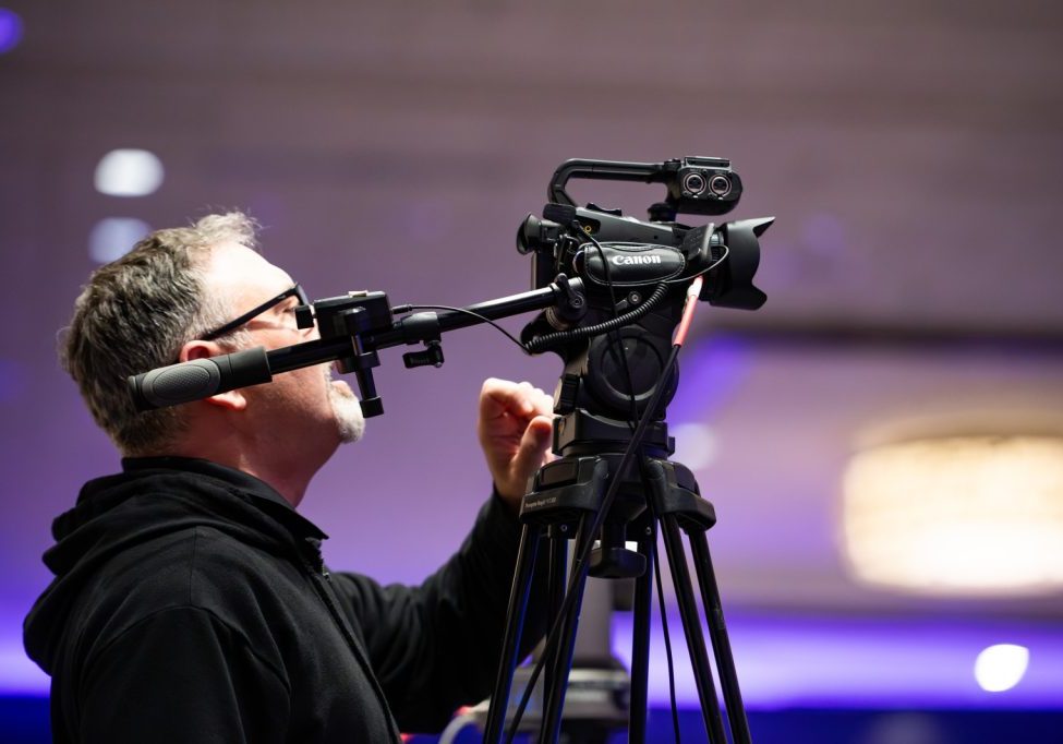 A man in glasses operates a Canon video camera mounted on a tripod indoors, adjusting the camera settings.