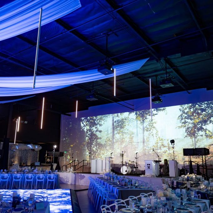 A decorated event space with blue lighting, draped white fabric on the ceiling, set tables, a chandelier, and a stage with musical instruments and a forest-themed backdrop.