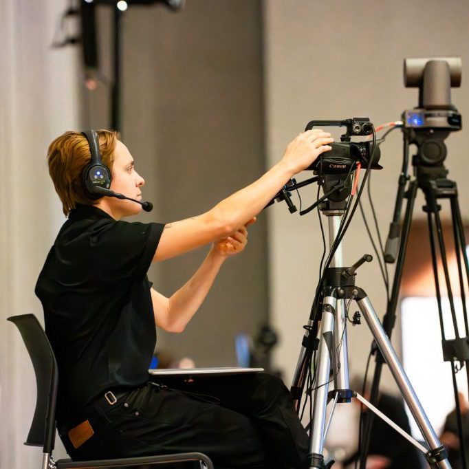 LB5_9874-Enhanced-NR (1) A person wearing headphones adjusts a video camera on a tripod during an indoor event.