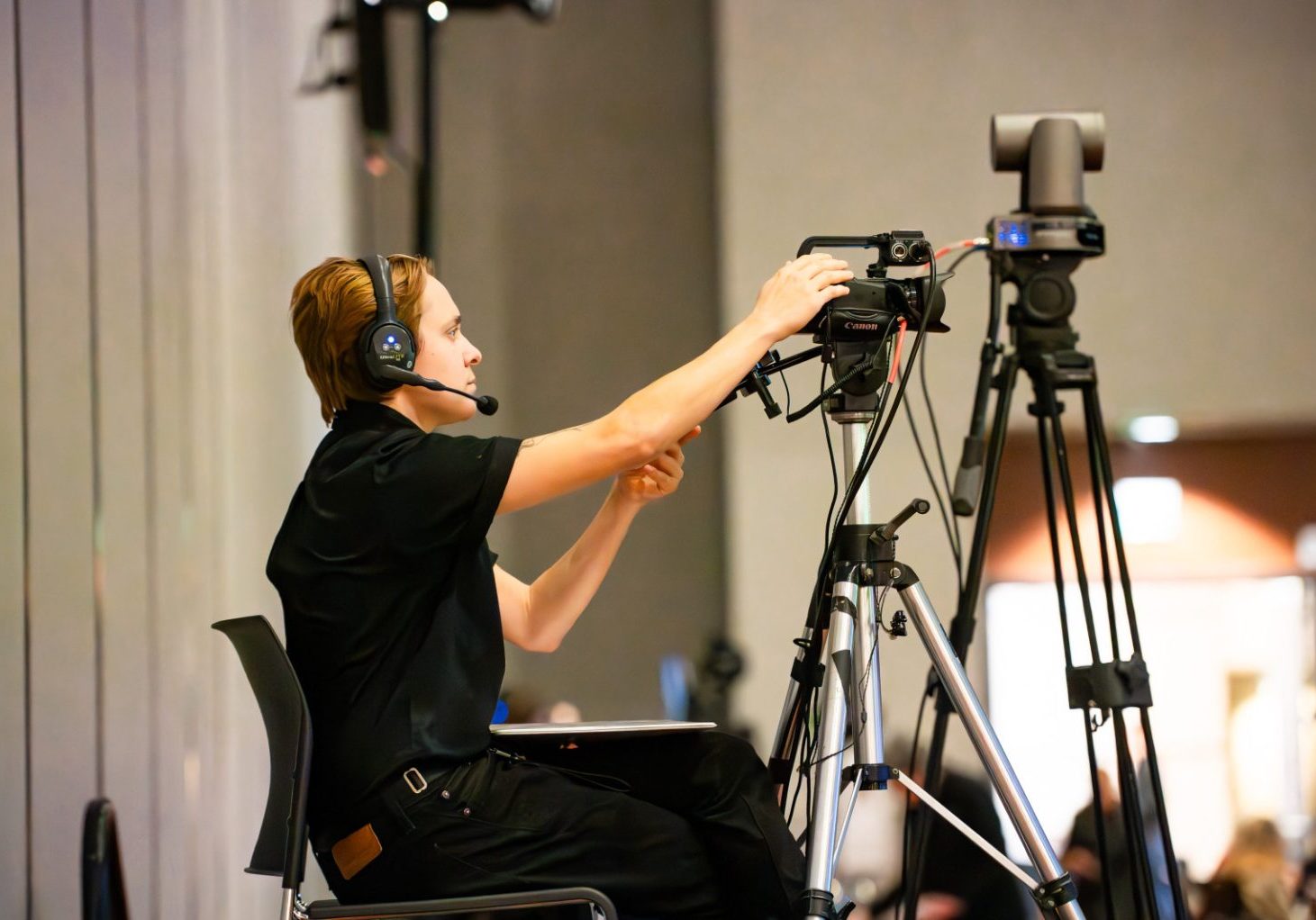 An Audio Visual Technician wearing a headset adjusts a video camera on a tripod in an indoor setting, possibly at an event or conference.